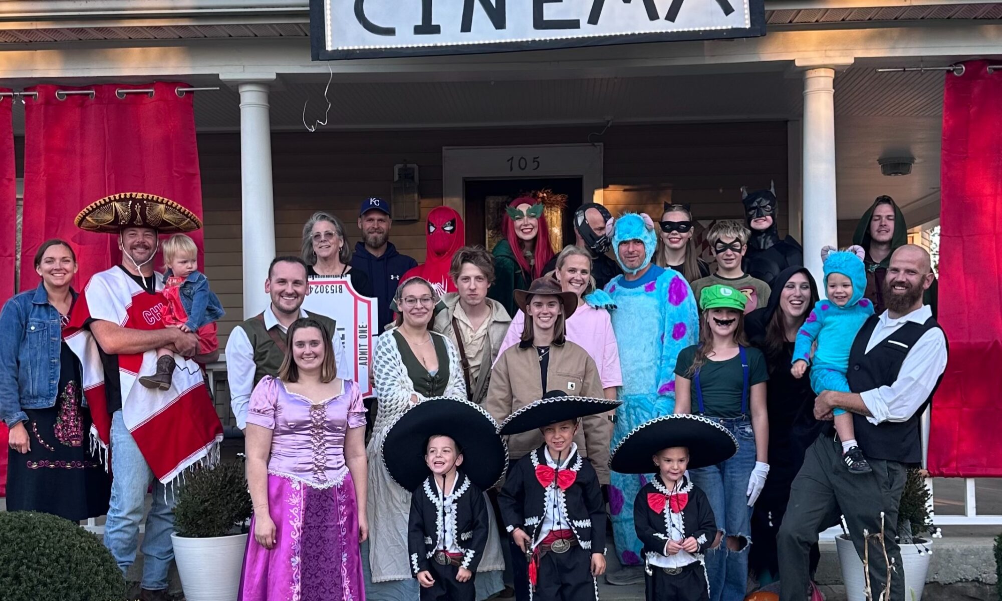 The large Doan Family, standing on a porch and down the porch stairs, to show off their Halloween costumes. Red curtains sit behind them along with a large sign that reads "Cinema" that greets trick-or-treaters when they come to the house.